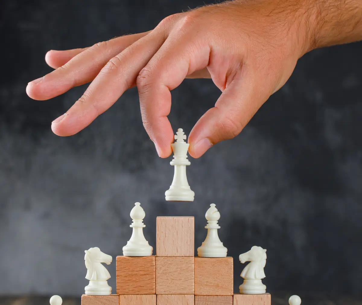 Een hand plaatst een witte schaakkoning bovenop een piramide van houten blokken en schaakstukken, wat symboliseert dat leiderschap of succes het resultaat is van een strategisch opgebouwde basis.